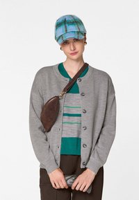 Grauer Cardigan mit Knopfschluss, über einem grün-grau gestreiften Hemd. Blaue Karokappe und braune Umhängetasche. Braune Hose vervollständigt das Outfit.