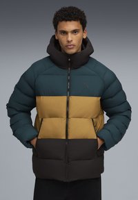 Homme portant une doudoune à blocs de couleurs teal, moutarde et noire, les mains dans les poches, se tenant devant un fond gris.