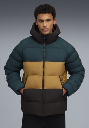Man in een teal, mosterdgeel en zwarte kleur geblokkeerde pufferjack, met zijn handen in zijn zakken, staand tegen een grijze achtergrond.