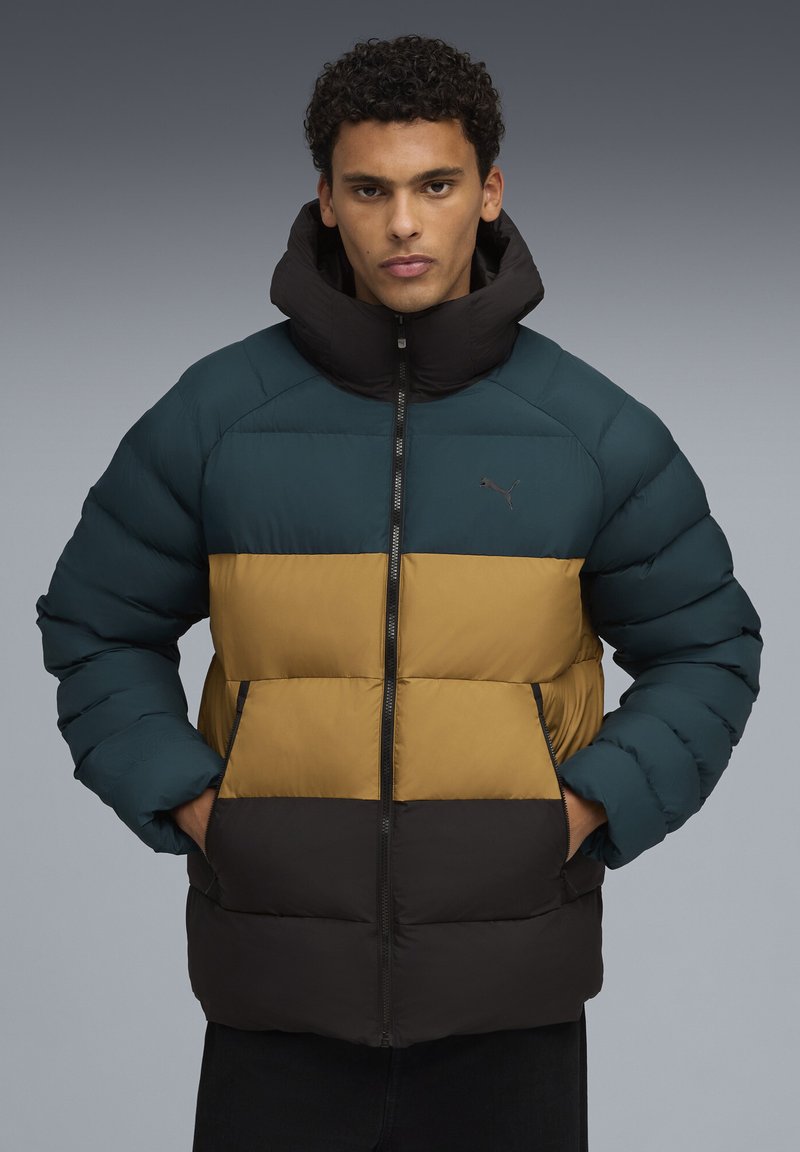 Homme portant une doudoune à blocs de couleurs teal, moutarde et noire, les mains dans les poches, se tenant devant un fond gris.