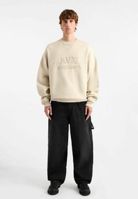 Felpa beige con testo in rilievo sul davanti, con scollo rotondo e vestibilità ampia. Abbinata a pantaloni neri a gamba larga e scarpe nere.