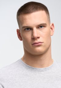 Pilkos spalvos marškinėliai su apvaliu kaklu, lygiu paviršiumi, prigludusio dizaino, ant modelio su trumpais plaukais ir neutralia išraiška, šviesiame fone.