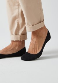 Ballet flats nere realizzate in tessuto morbido con un'area della punta testurizzata, abbinate a pantaloni beige arrotolati, che mettono in evidenza dettagli di caviglia e piede.