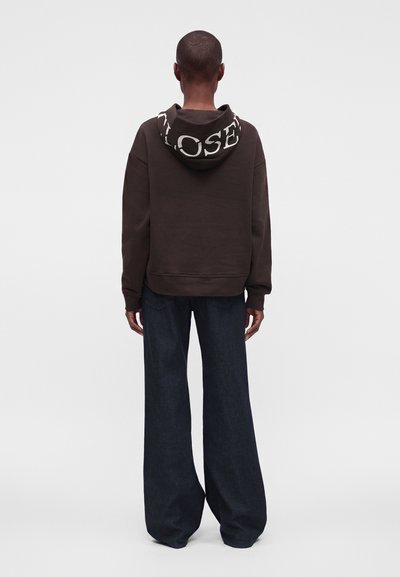 Personne de dos, portant un sweat à capuche sombre avec de grandes lettres blanches sur la capuche et un pantalon large et foncé, debout devant un fond uni.