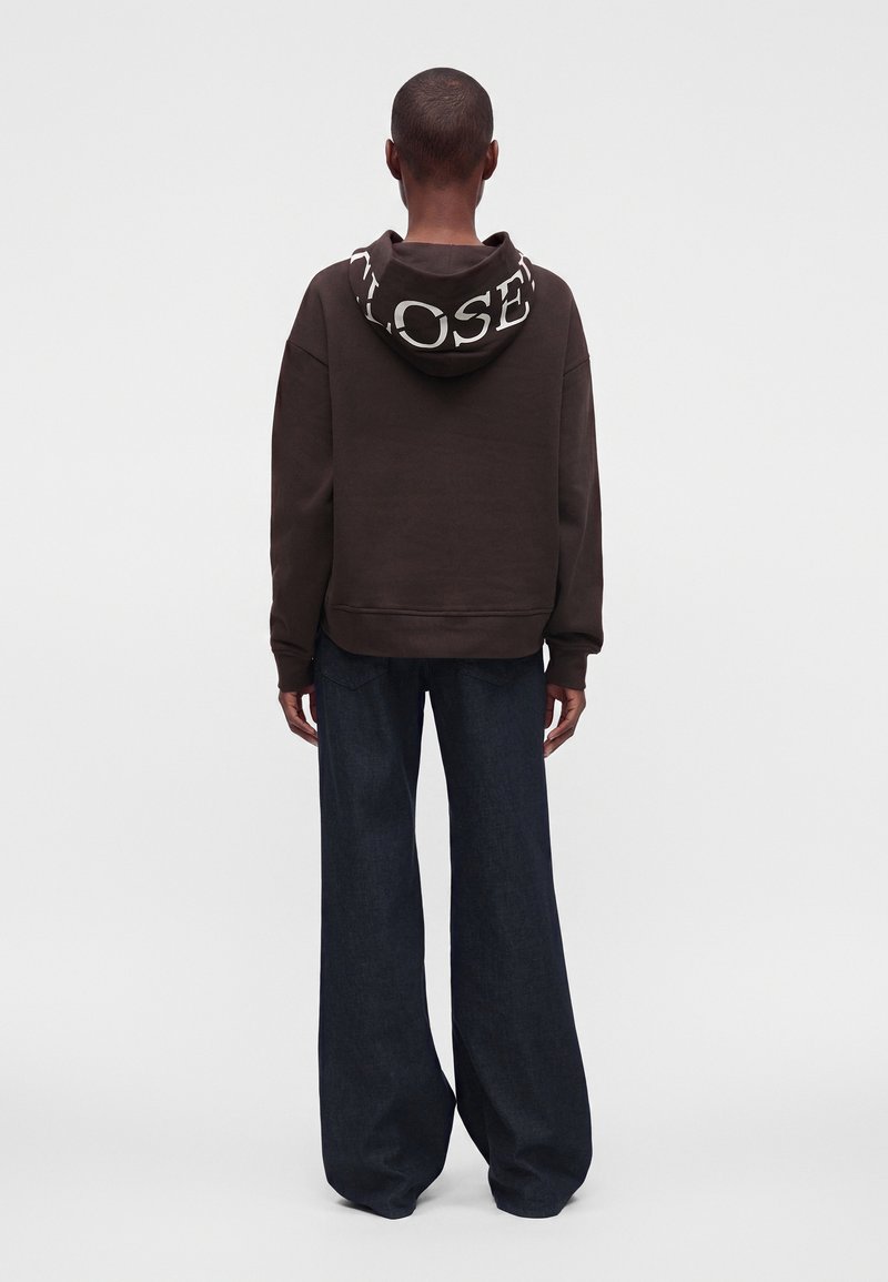 Personne de dos, portant un sweat à capuche sombre avec de grandes lettres blanches sur la capuche et un pantalon large et foncé, debout devant un fond uni.