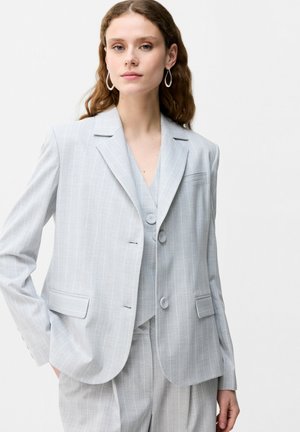 Kobieta o falowanych brązowych włosach, ubrana w jasnoszary, cienko prążkowany, dopasowany marynarkę, pasującą kamizelkę i spodnie, stojąca z ręką w kieszeni.