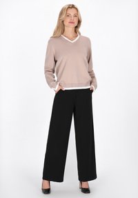 Pulover bej deschis cu decolteu în V și bordură albă, asortat cu pantaloni negri largi. Modelul are mâinile în buzunare și poartă pantofi cu toc negri.