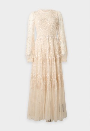 Femme aux longs cheveux foncés portant une robe maxi en dentelle beige transparente à manches longues avec broderie florale, debout sur un fond blanc.