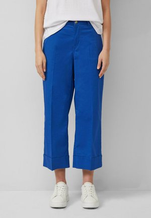 Osoba ubrana w jasnoniebieskie, skrócone spodnie o szerokich nogawkach, białe sportowe buty i białą bluzkę wpuszczoną do środka, stojąca na tle jednolitej ściany.