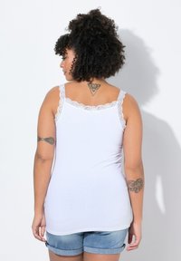Top bianco senza maniche con pizzo ai lati delle spalle, design aderente e texture liscia. Indossato con pantaloncini di jeans, che mettono in mostra tatuaggi sulla schiena.