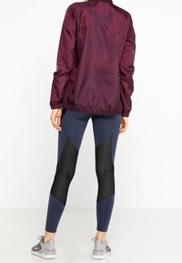 Rompevientos burdeos con cuello alto, leggings navy con paneles de malla y zapatillas deportivas grises con acentos rosas.