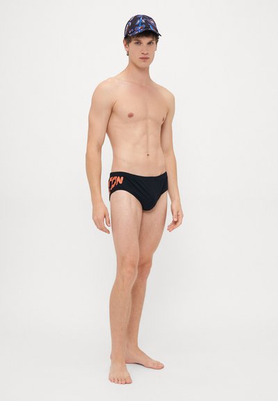 Junger Mann steht barfuß da, trägt eine schwarze Badehose mit orangefarbener Schrift und eine blaue gemusterte Kappe vor einem weißen Hintergrund.