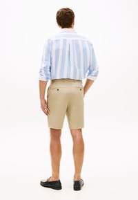 Άνδρας με την πλάτη γυρισμένη φοράει πουκάμισο με σιέλ και λευκές ρίγες, μπεζ σορτς και μαύρα μοκασίνια, στέκεται μπροστά σε απλό λευκό φόντο.