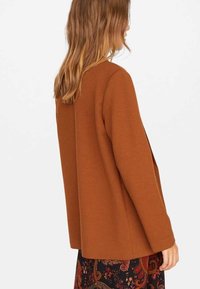 Hnědý oversized blazer vyrobený z texturované látky, s rovnými linemi a otevřeným předním dílem, doplněný vzorovanou sukní v tmavých barvách.