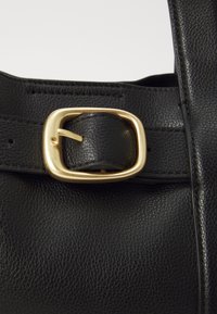 Schwarze Lederhandtasche mit strukturiertem Oberflächen. Verfügt über eine goldene rechteckige Schnalle und einen Trageriemen. Nahaufnahme der Schnalle und des Materials sichtbar.