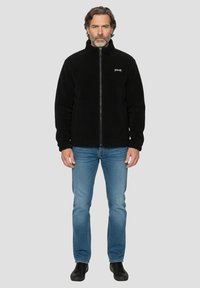 Schwarze Fleecejacke mit Frontreißverschluss und hohem Kragen, kombiniert mit blauen Jeans und schwarzen Schuhen. Die Jacke hat eine weiche Textur und dezentes Branding.