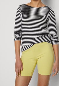 Kobieta ubrana w czarną i białą paskowaną bluzkę z długim rękawem, z podwiniętymi rękawami, oraz żółte dopasowane szorty do kolan.