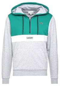 Sudadera con capucha de sección superior verde, franja horizontal blanca, sección inferior gris, cremallera frontal, bolsillo tipo canguro y logo de Lacoste en el pecho.