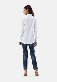 Weißes Langarmhemd mit abgerundetem Saum, geknöpften Manschetten und klassischem Kragen, kombiniert mit blauen Jeans und beigen High-Heels.