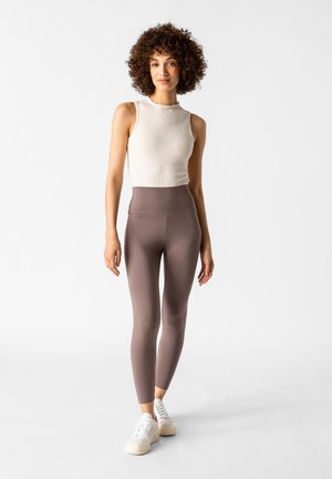 Leichtes, geripptes weißes Tanktop mit Rundhalsausschnitt, kombiniert mit hoch taillierten, figurnahen, dunkelbraunen Leggings und weißen Turnschuhen.
