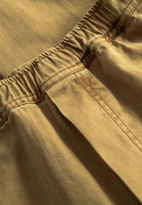 Beiger Fischgrätstoff mit elastischem Bund. Das Material hat eine glatte Textur mit dezentem Glanz und Falten für Drapierungen.