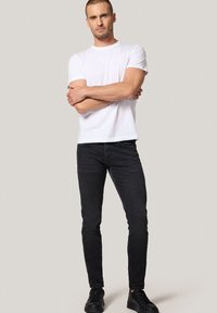 Mężczyzna z krótkimi włosami, ubrany w białą koszulkę, czarne dopasowane dżinsy i czarne sneakersy, stojący z założonymi rękami na beżowym tle.