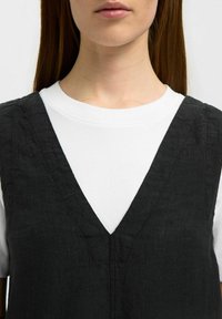 Zwarte linnen jurk met een V-hals ontwerp, gelaagd over een wit t-shirt. Zichtbare stiksels op het kledingstuk. Over het geheel een gladde textuur.