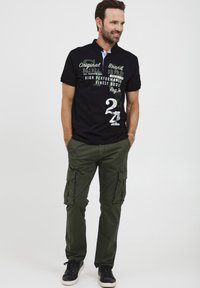 Melna polo krekls ar baltām un zaļām tekstu grafiskām detaļām un gaiši zilu apkakli, kombinēts ar olīvu krāsas cargo biksēm un melniem sporta apaviem.