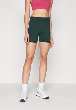 Frau trägt dunkelgrüne enganliegende Shorts, einen pinken Sport-BH, weiße Socken und weiße Sportschuhe, steht vor einem einfarbigen hellen Hintergrund.