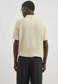 Beige Poloshirt aus strukturiertem Stoff, mit kurzen Ärmeln und einer Rückenpasse, kombiniert mit schwarzen Hosen mit Taschen.