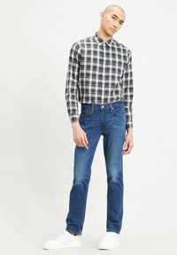 Camicia a quadri in verde, blu navy e bianco; abbinata a jeans in denim blu; modello che indossa sneaker bianche, mani posizionate ai lati.