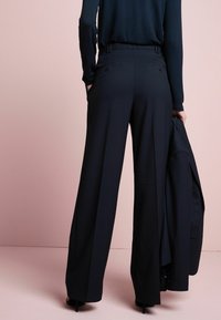Sorte brede bukser med en glat tekstur, som har baglommer og bæltestropper, parret med en matchende blazer over armen.