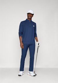 Námořnická modrá golfová mikina s dlouhým rukávem a zipem, v kombinaci se slim-fit kalhotami a bílými golfovými botami, držící golfovou hůl.