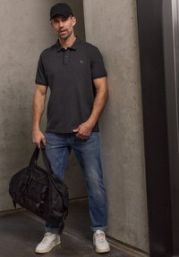 Homme en polo noir, jean bleu et baskets blanches tenant un sac de sport noir, debout contre un mur en béton près d'un ascenseur.