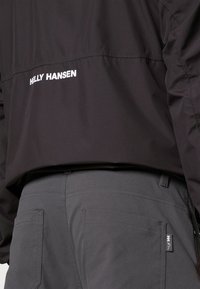 Schwarze Helly Hansen Jacke mit einem weißen Logo, kombiniert mit grauen Hosen, die minimale Nähte und zwei Gesäßtaschen aufweisen.