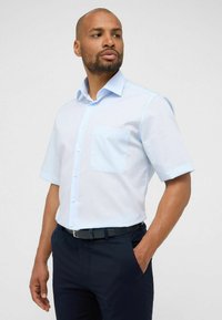 Camicia azzurra a maniche corte con bottone frontale, taschino sul petto e texture uniforme, abbinata a pantaloni scuri e una cintura nera.