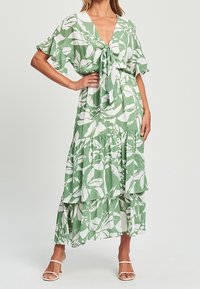 Vestido verde y blanco con estampado floral, escote en V, lazo delantero y falda con volantes en capas. Mangas cortas y acampanadas de tela ligera.
