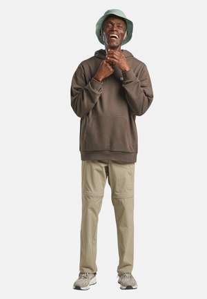 Lächelnder Mann mit grünem Anglerhut, braunem Kapuzenpullover, beiger Hose und Turnschuhen, der die Kordeln seines Kapuzenpullover vor einem einfarbigen weißen Hintergrund anpasst.