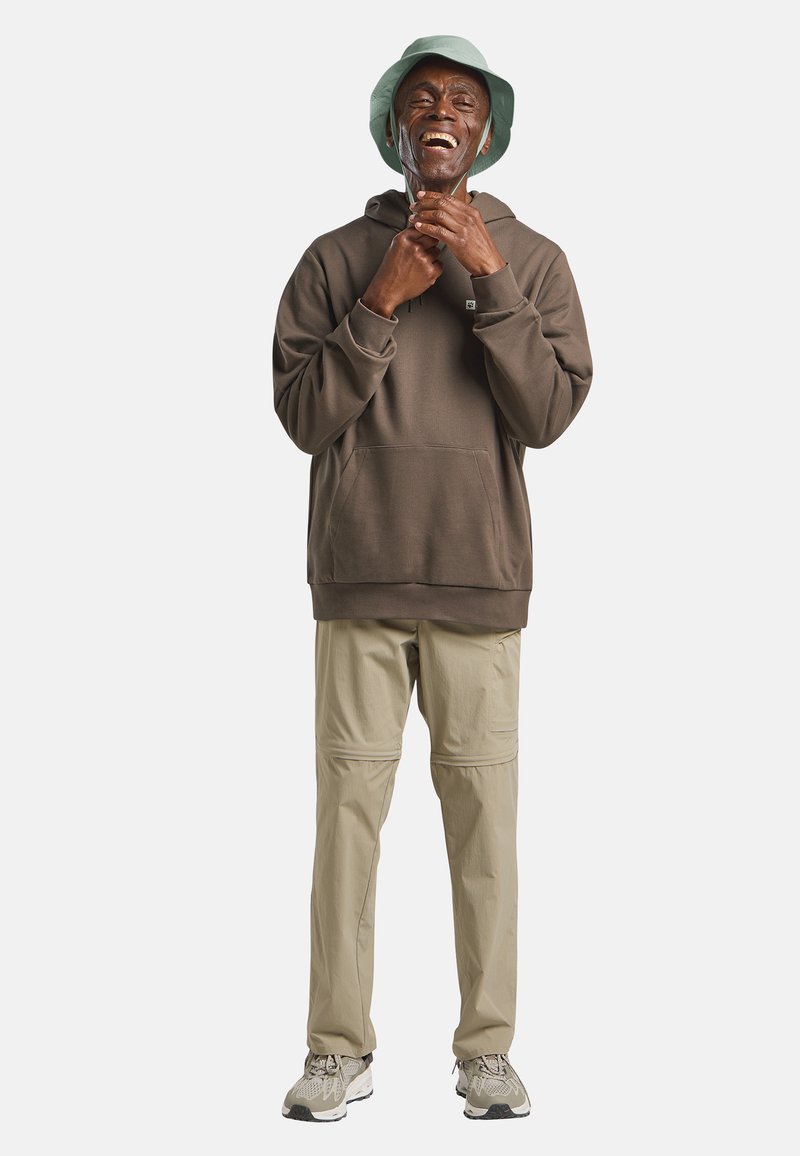 Glimlachende man met een groene vissershoed, een bruine hoodie, beige broek en sneakers die de trekkoorden van zijn hoodie aanpast tegen een effen witte achtergrond.