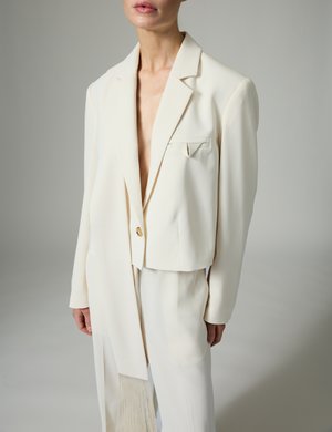 Person, die einen cremefarbenen asymmetrischen Blazer mit einem Knopf sowie passende hoch taillierte Hosen vor neutralem Hintergrund trägt.