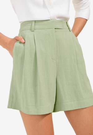 Vrouw die lichtgroene geplooide shorts met hoge taille en zakken draagt, gecombineerd met een wit shirt met lange mouwen dat is ingestopt.