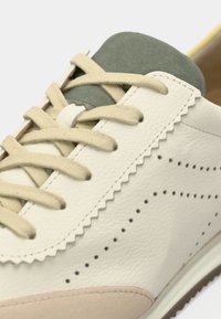 Αθλητικό παπούτσι με λευκό δερμάτινο επάνω μέρος, γλώσσα από πράσινο σουέτ και κορδόνια σε κρεμ απόχρωση. Διαθέτει ανάγλυφες λεπτομέρειες με κυματιστές άκρες και διάτρητες διακοσμήσεις. Καφέ σόλα.