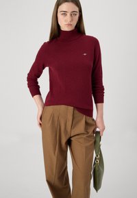 Burgunderfarget rullekragesweater laget av mykt stoff, sammen med høyt liv brune bukser. En grønn håndveske holdes i den ene hånden.