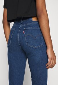 Vue arrière d'une personne portant un jean taille haute bleu avec des poches arrière et un haut court noir, le bras détendu le long du corps.