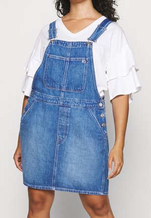 Woman wearing a blue denim overall dress with front pocket buttons, paired with a white short-sleeve ruffled blouse.