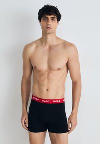 Schwarze Boxershorts mit einem roten elastischen Bund, auf dem das Logo "HUGO" deutlich sichtbar ist. Der Stoff wirkt glatt und körperbetont.