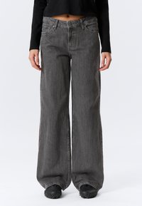 Graue Denim-Jeans mit weitem Bein und used Look, ausgestattet mit vorderen Taschen und Knopfverschluss. Kombiniert mit einem schwarzen, bauchfreien Top.