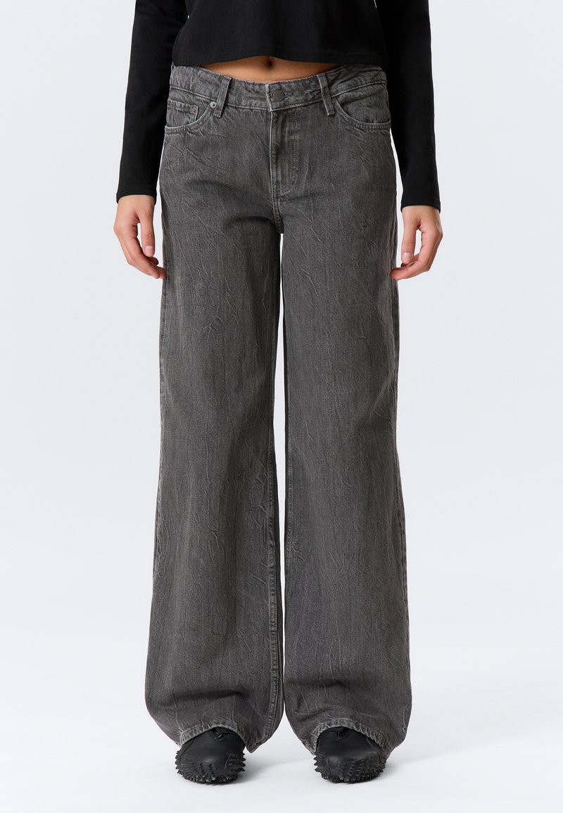 Graue Denim-Jeans mit weitem Bein und used Look, ausgestattet mit vorderen Taschen und Knopfverschluss. Kombiniert mit einem schwarzen, bauchfreien Top.