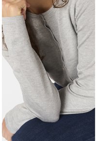 Femme portant un pull côtelé gris clair à manches longues avec des boutons, reposant sa tête sur un poing fermé, assise avec un pantalon foncé.