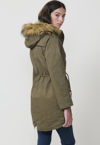 Olíva zöld parkajacket műszőrmével bélelt kapucnival, húzózsinórral ellátott derékrésszel és hosszú ujjal. Egyenes vonalú fazon és enyhén ívelt alj jellemzi.